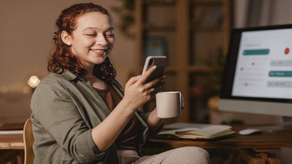 mulher em casa usando smartphone na frente computador enquanto toma cafe